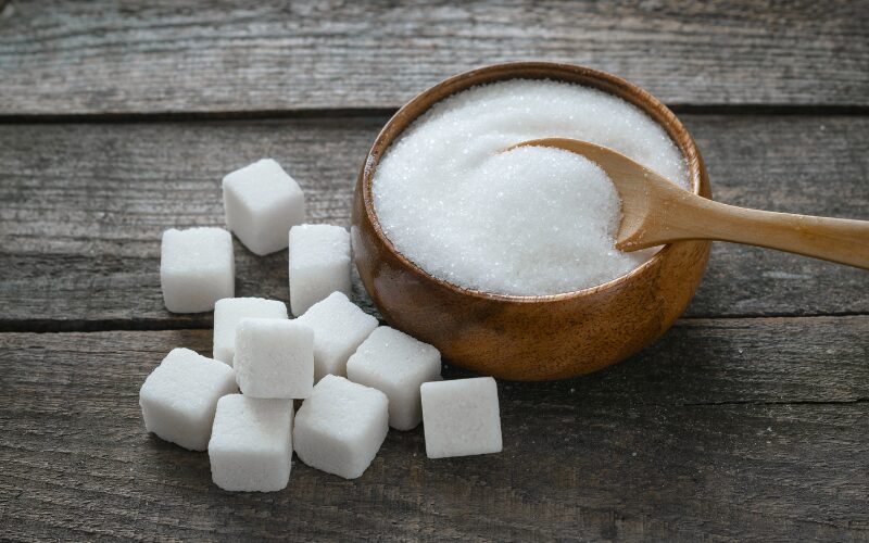 Pile of sugar cubes beside bowl of refined sugar with wooden spoon, showing sugar blues impact and ways to reduce intake.
