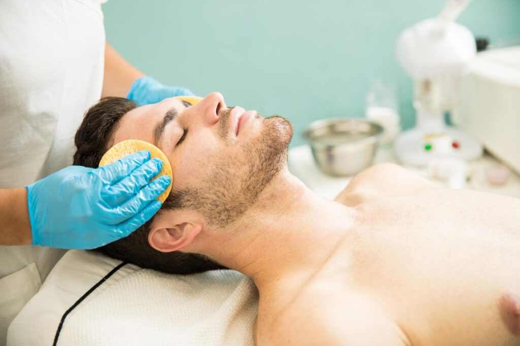 Therapist cleansing a man's face with a sponge, illustrating key steps in male wellness treatments.