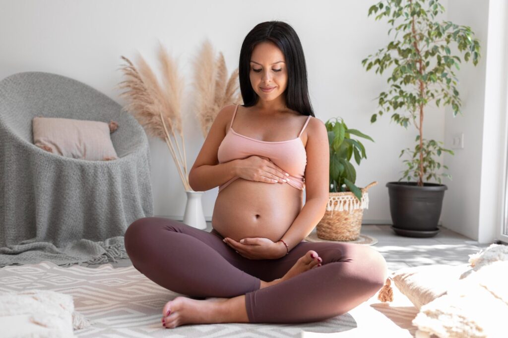  A pregnant woman at Beauty Kliniek sits cross-legged and gently holds her belly during a wellness session. 
