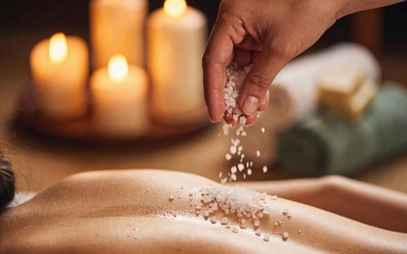 Therapist sprinkling sea salt on woman's back during detox scrub in candlelit spa for improved skin clarity and energy.