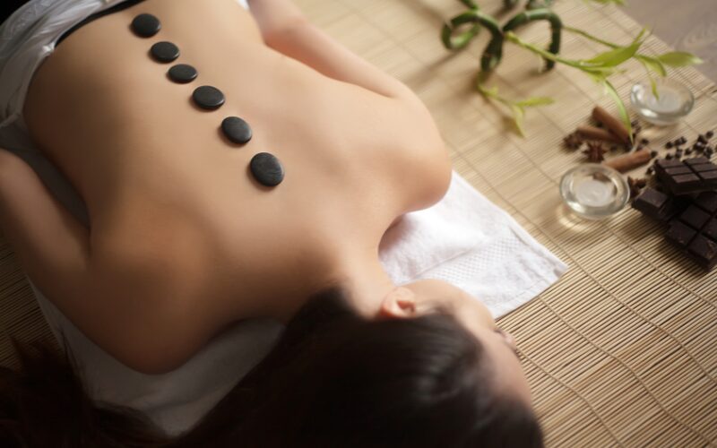 Woman relaxing with hot stones aligned on back, surrounded by bamboo and herbs, enhancing detox pathways for better energy and skin.