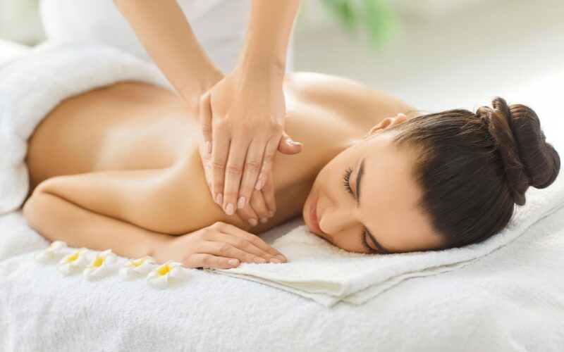 Therapist applying mud mask to woman's back during spa session with natural ingredients tray, promoting regular professional visits.